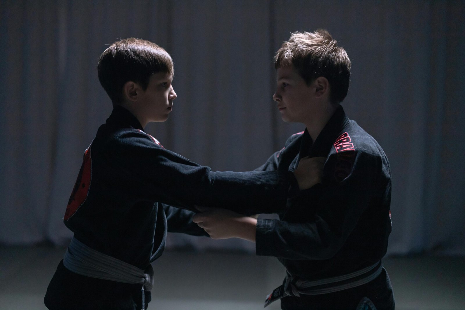 Two young boys in jiu-jitsu uniforms sparring indoors under low light.