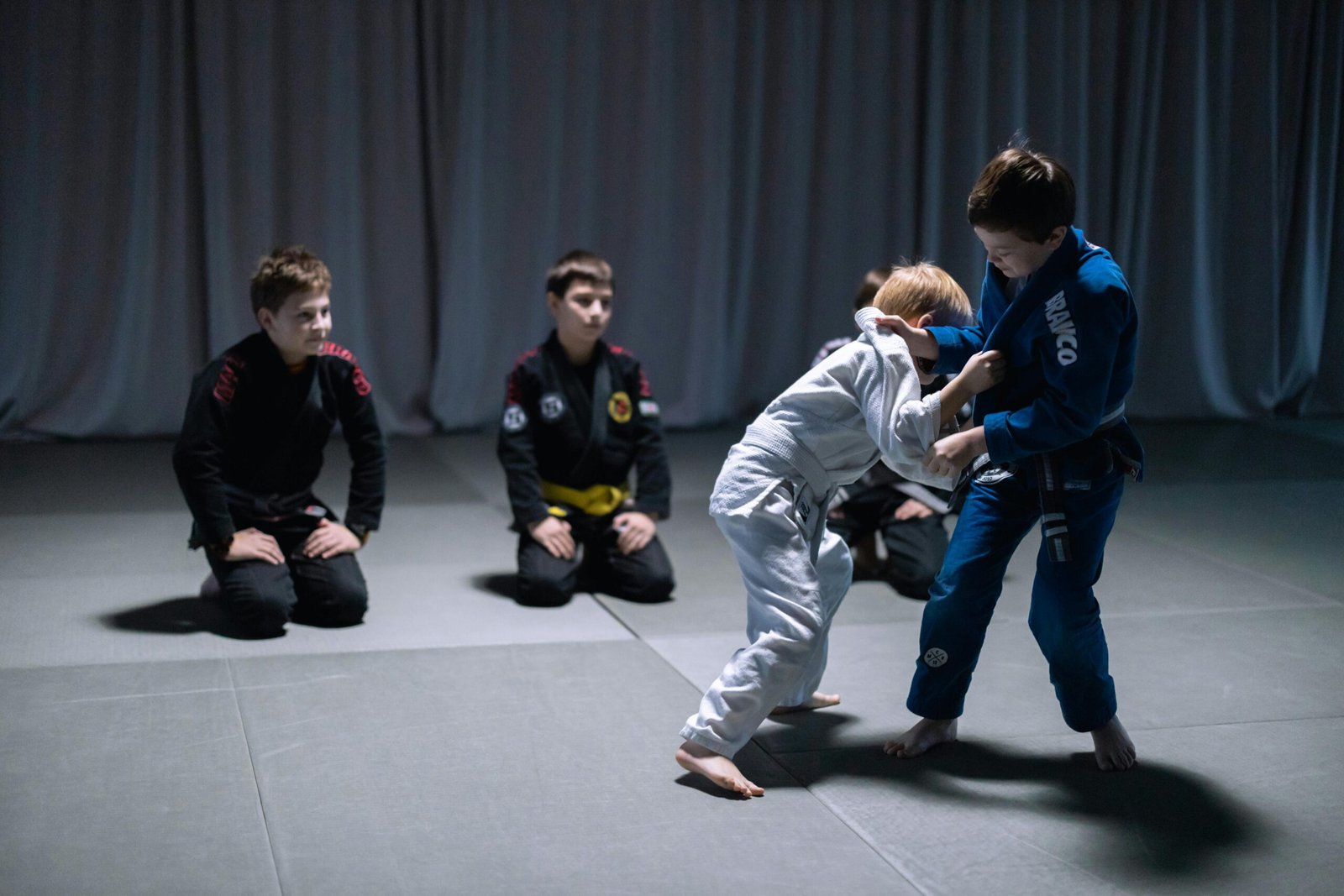 Children practicing jiu jitsu in a gym setting, wearing gi uniforms, during sparring session.