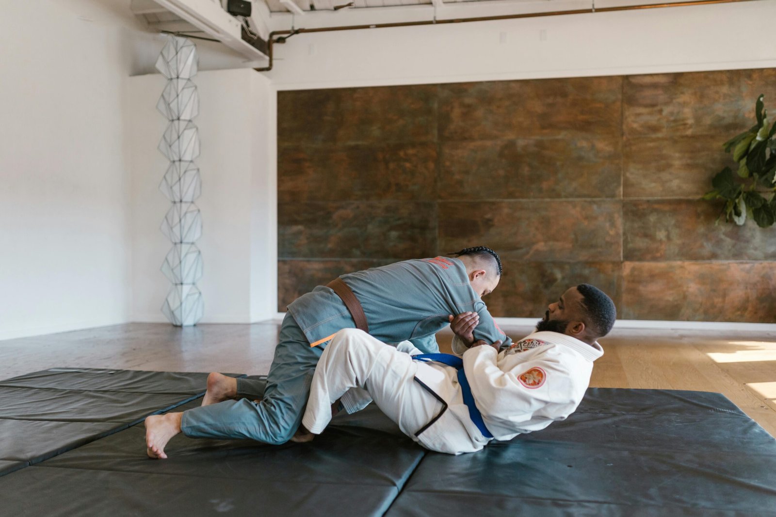 Two men in martial arts uniforms practicing Brazilian Jiu-Jitsu on mats indoors.
